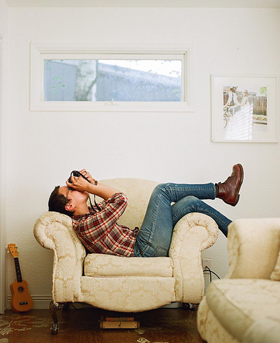 Brian Adams About Photo A man lying longways on a chair rests his back against one arm and drapes his legs over the other. He holds a camera to his face.
