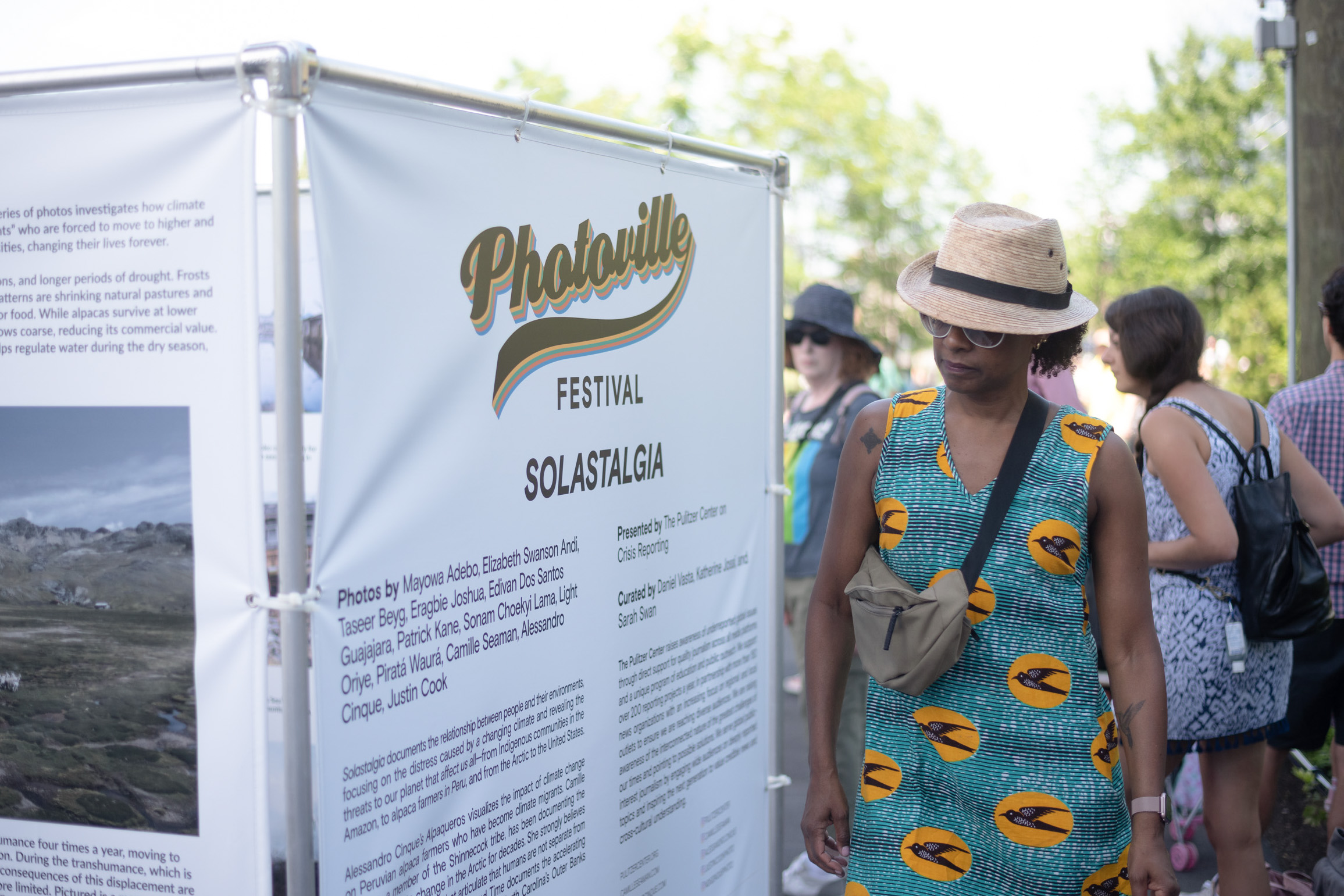 A woman in a sunhat and dress reads a Photoville poster