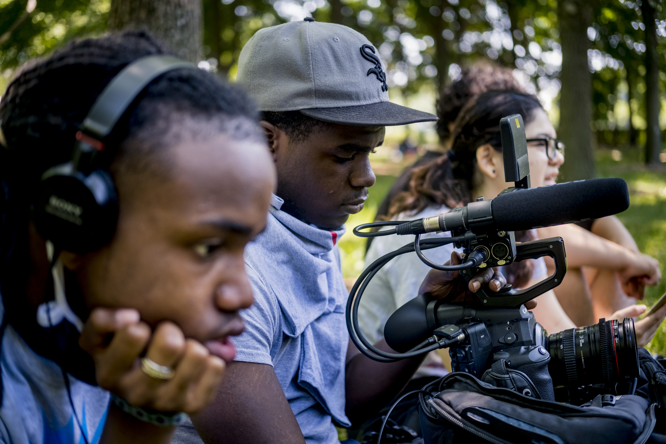 Four students work on creating a documentary. In the foreground, a student wears headphones to listen to audio they are recording. Another student uses a video camera to film. In the background, two students sit and watch the scene.