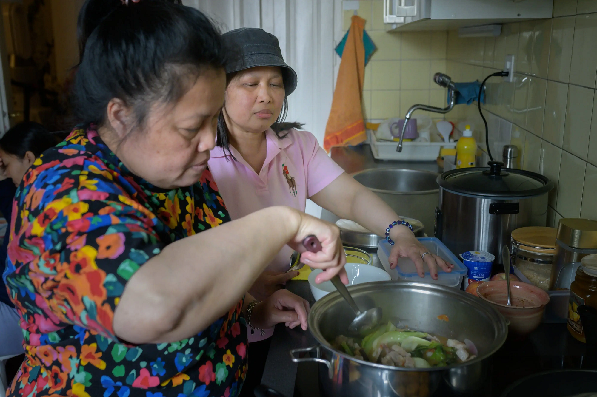 2023.05.07_GeorgiaAndFamily_32_Z628180_print-1684934412-min.png Two women are in a kitchen. One stirs a pot on the stove with meat and cabbage, and the other looks on in support.