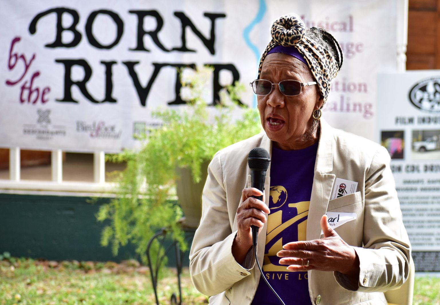 A woman talks into a microphone in front of a mural that reads &quot;Born by the River.&quot;