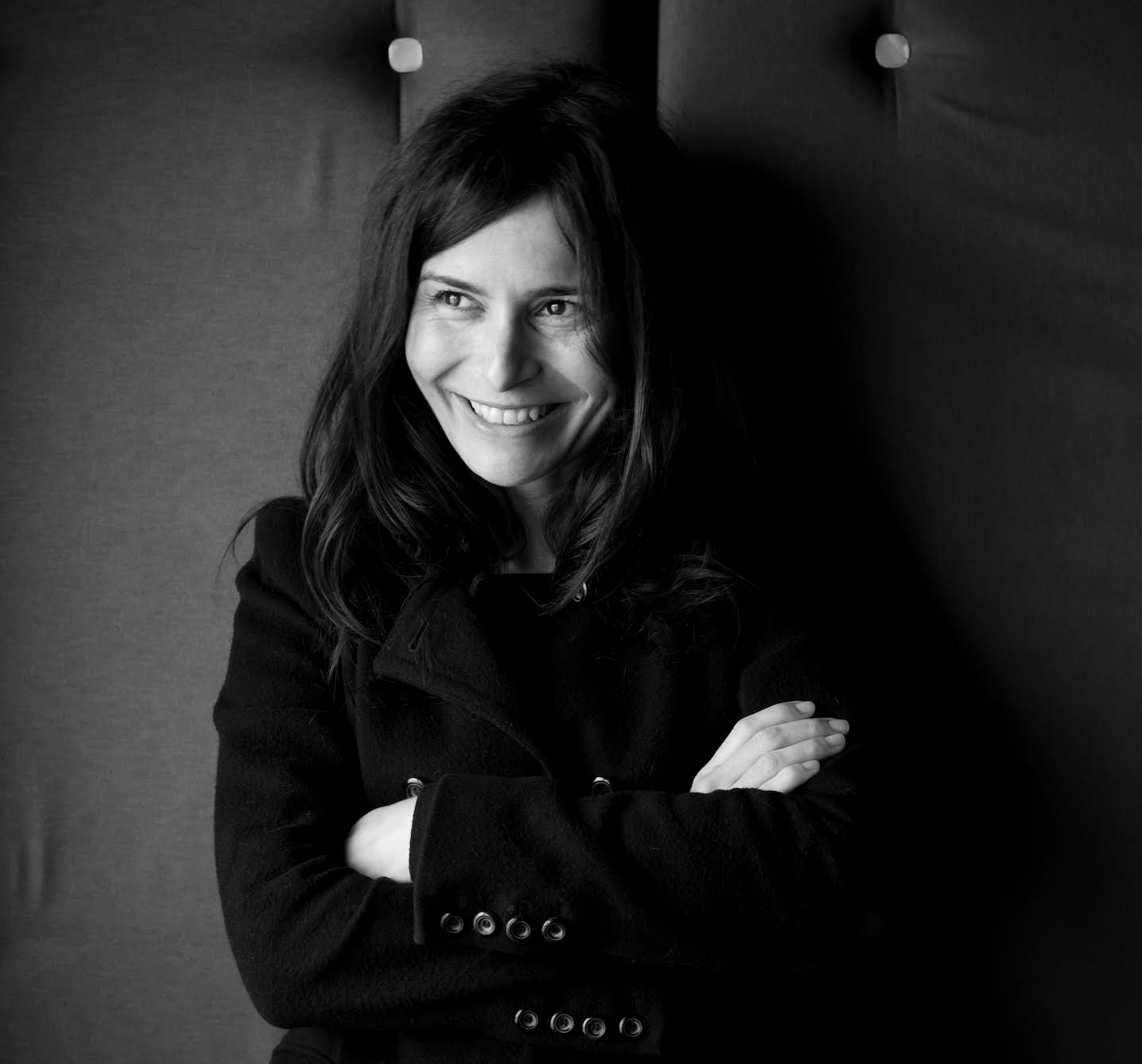 A black and white photo of a woman with long dark hair sitting down with her arms crossed, wearing a black jacket