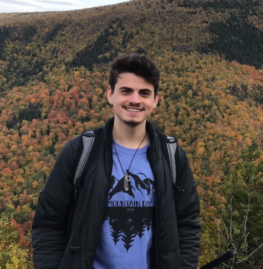 Man stands in black jacket and blue shirt in front of forest