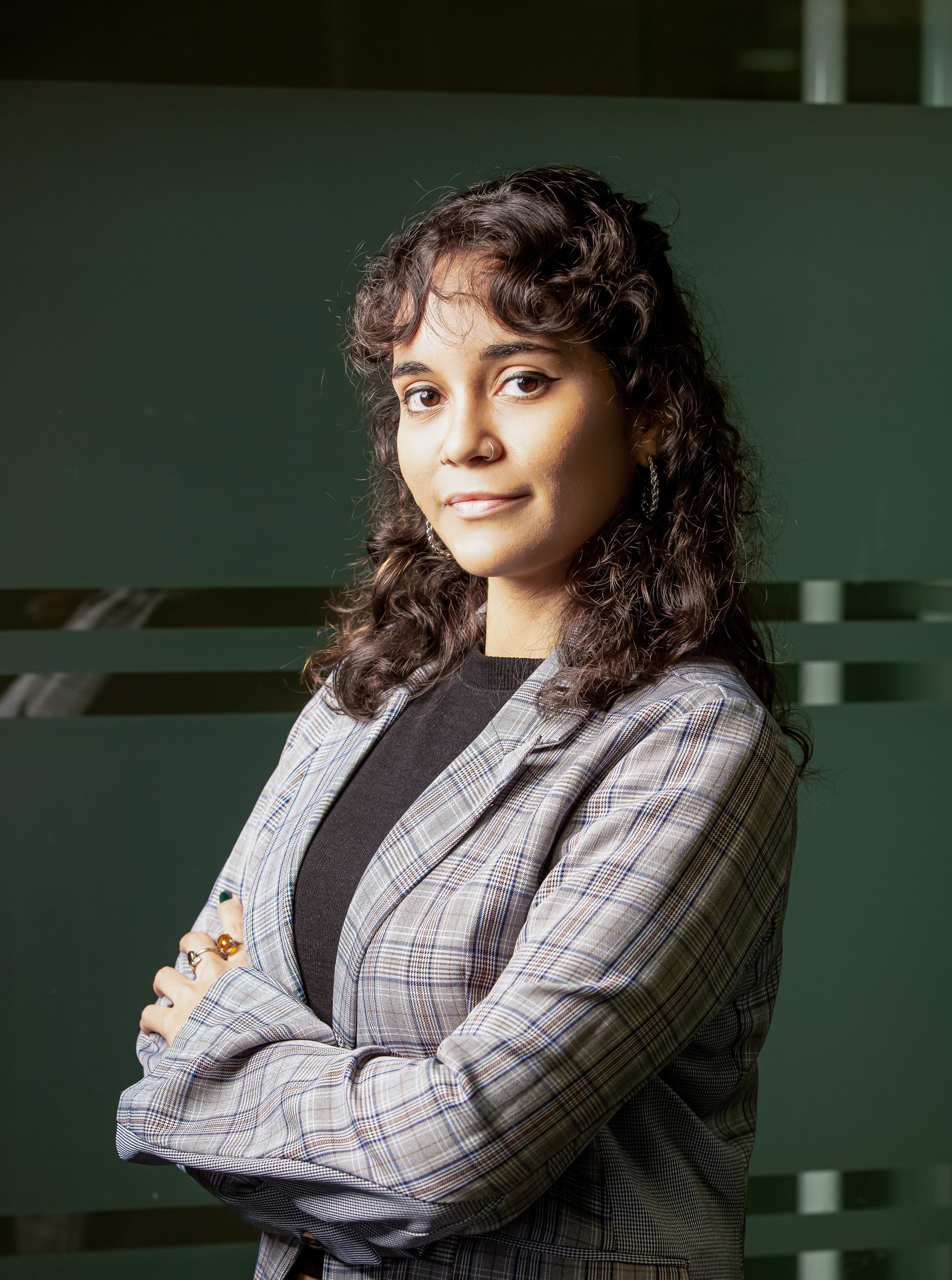 A woman with long curly hair stands against a dark background, arms crossed