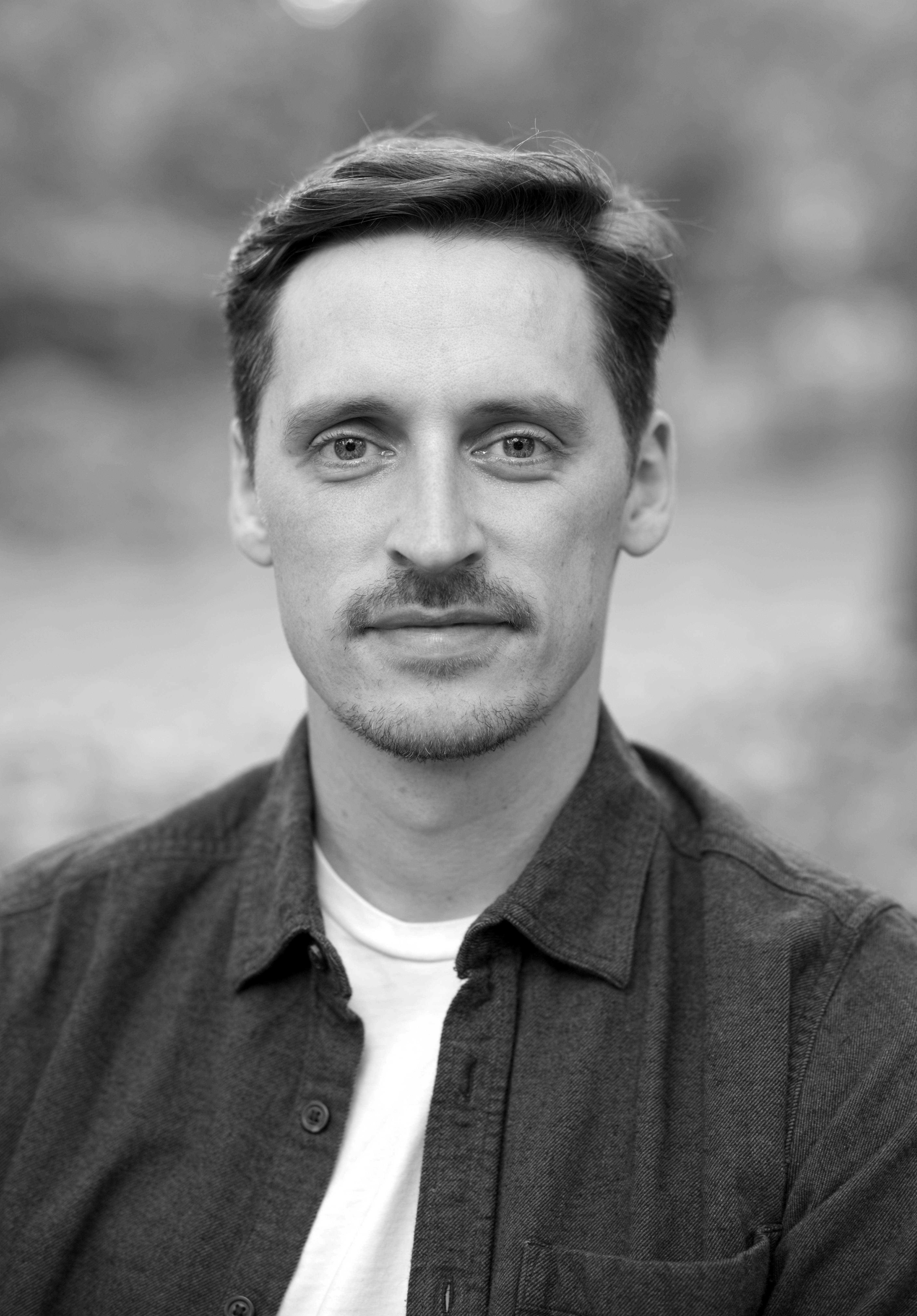 A black and white photo of a man with short dark hair wearing a white tshirt and a dark button down