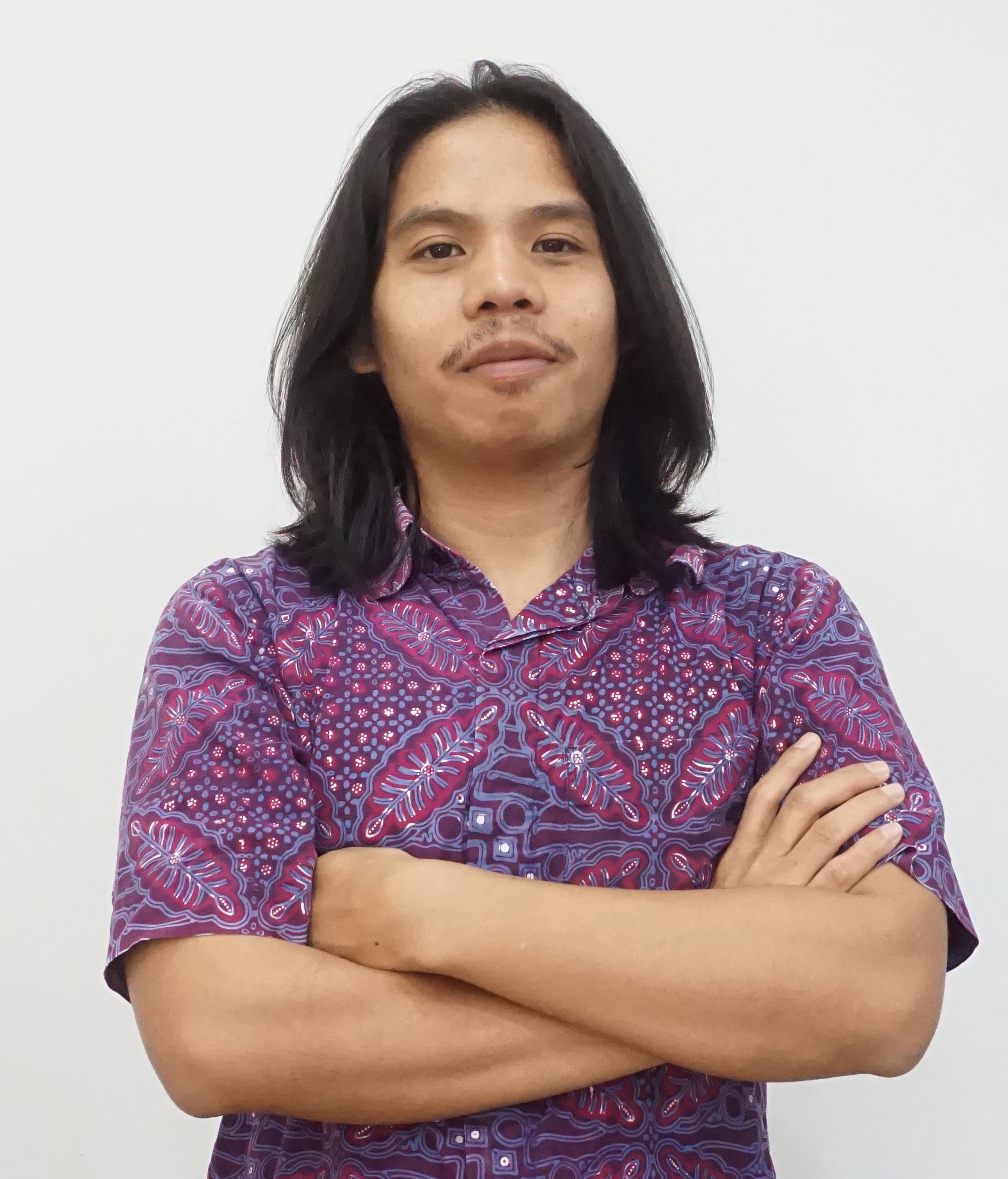 A man with long dark hair and wearing a purple shirt folds his arms
