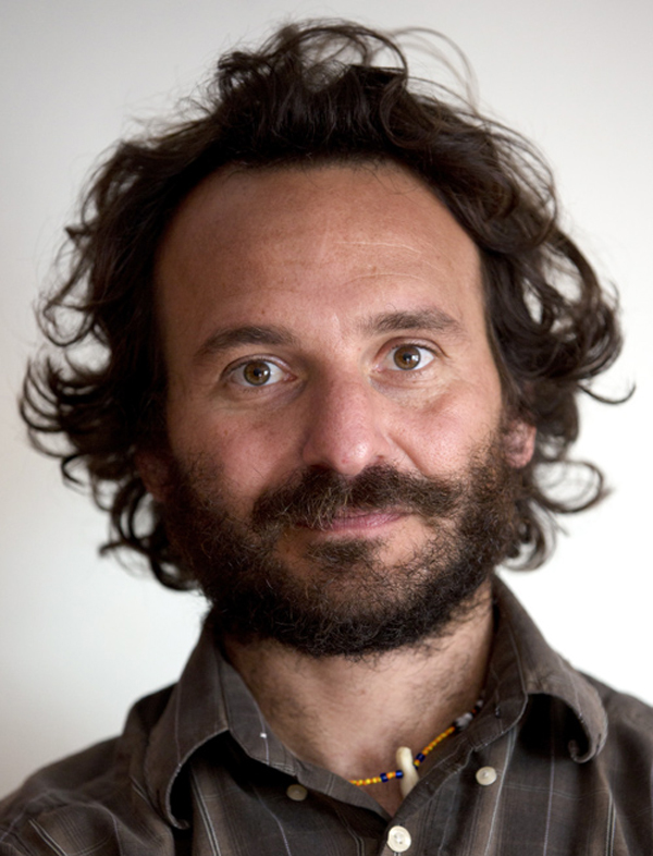 A man with curly brown hair and facial hair wears a brown button down
