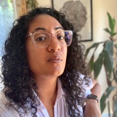 A woman with curly hair and glasses looks at the camera