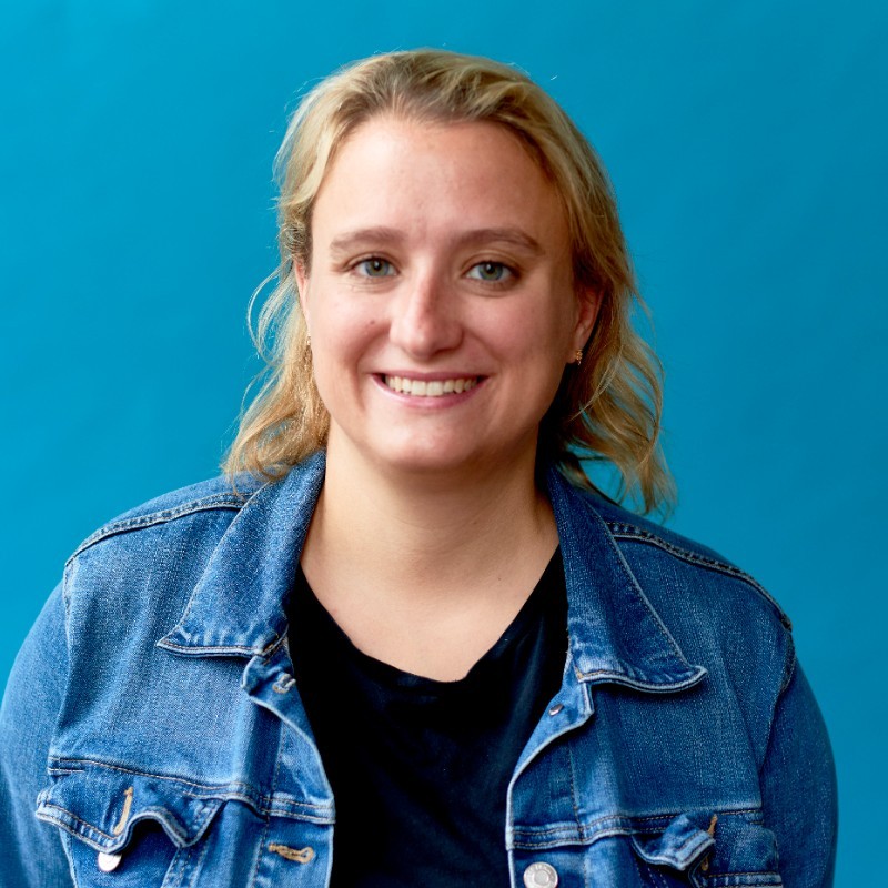A blonde woman wears a jean jacket against a blue background