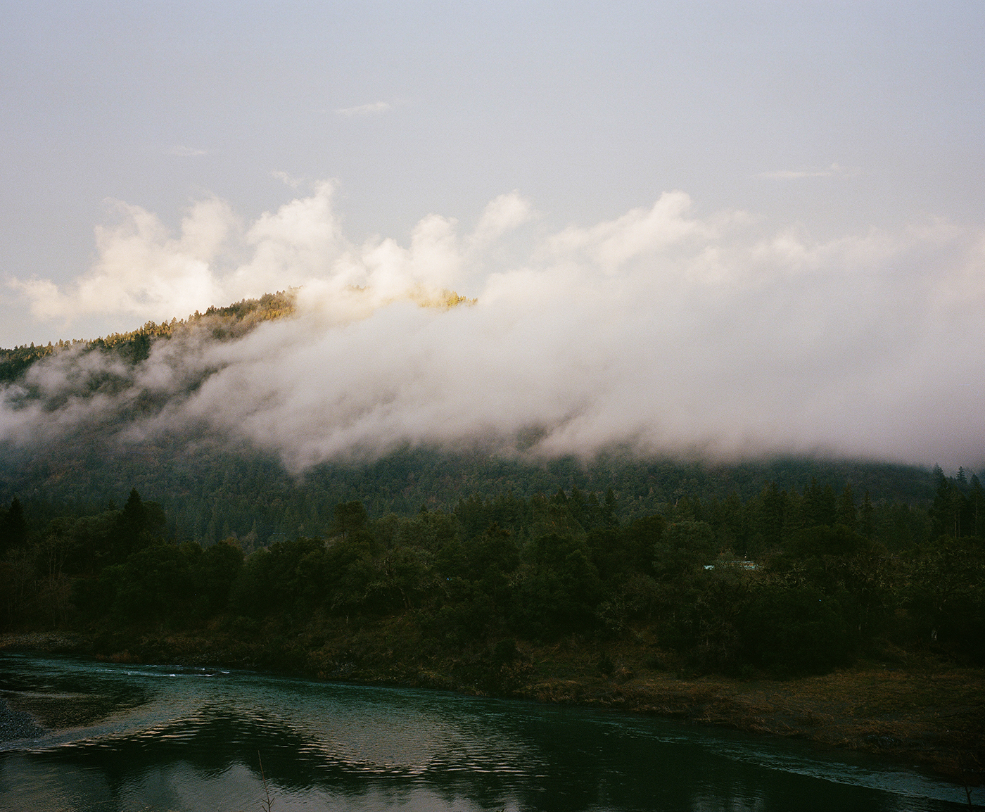 Clouds enshroud a green mountain, with a river at its base