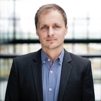 A man with short hair wearing a blue button down with a black blazer