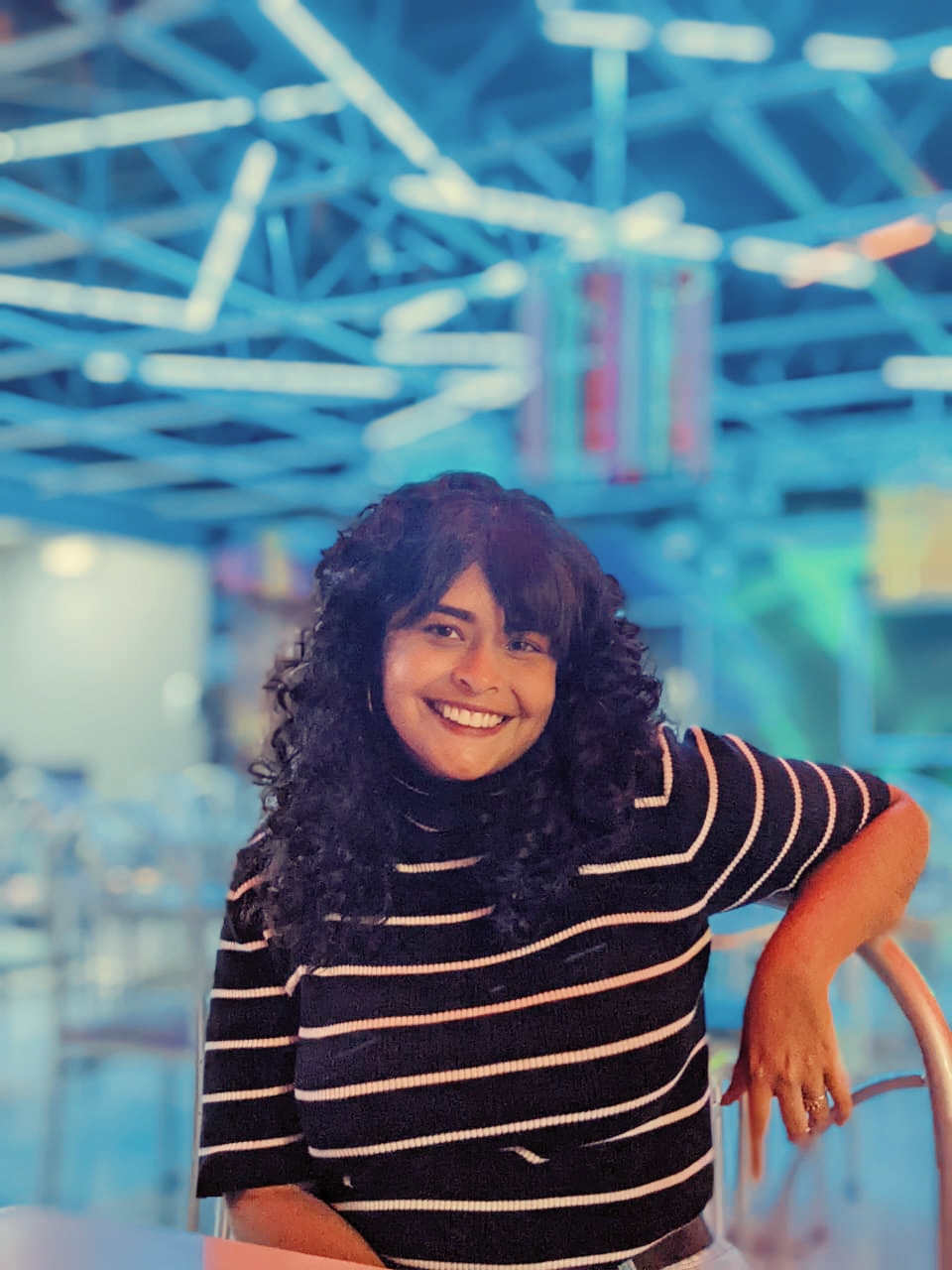 A woman with dark curly hair wearing a striped shirt against a bright blue-lit background
