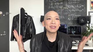 A person with a bald head wearing a leather jacket sits at a desk and gestures with both hands