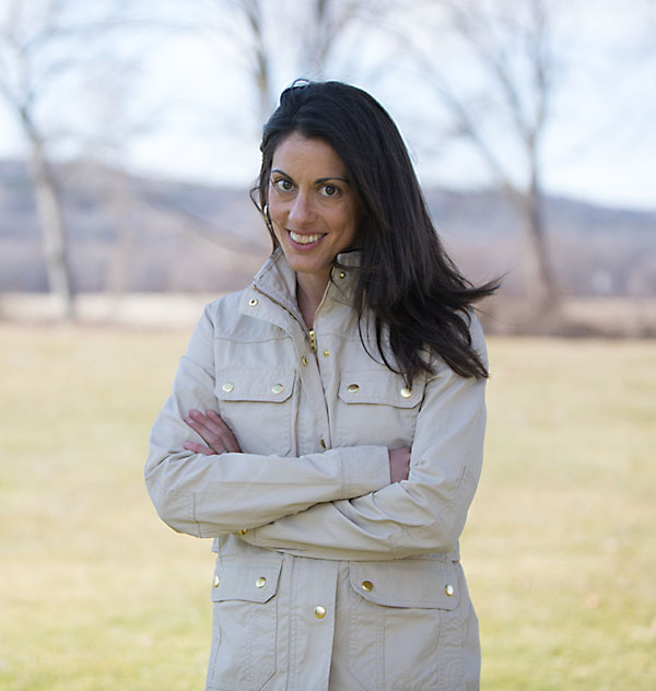 A woman with long dark hair stands in a field wearing a white coat with her arms crossed