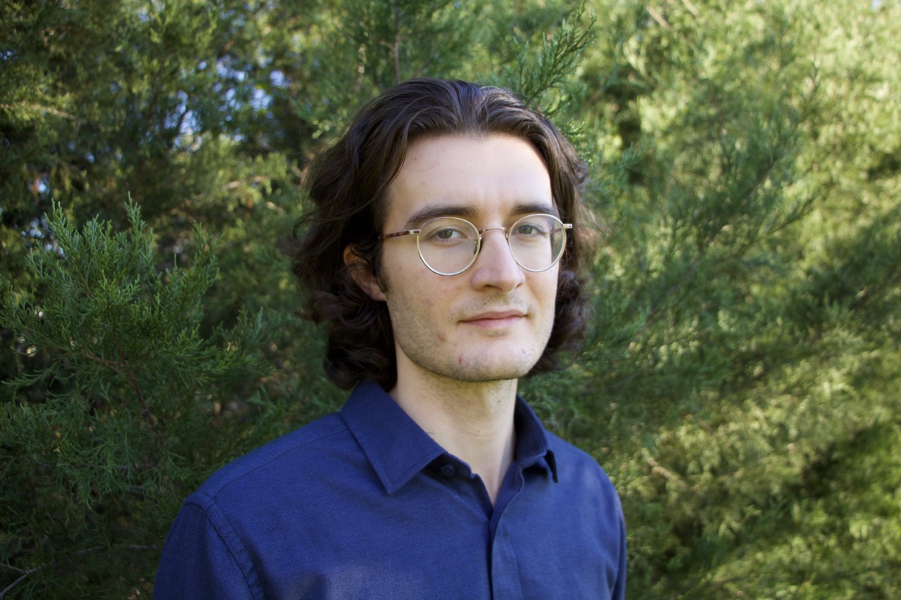 A man with dark hair and glasses wearing a blue button-down against a green leafy background