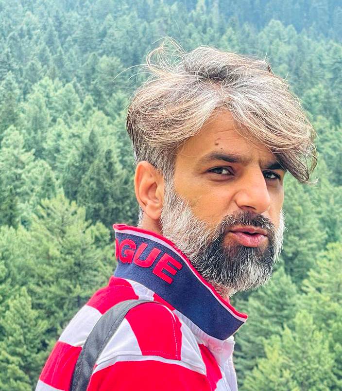 A man with grey hair and a beard wears a red and white striped shirt against a wooded background