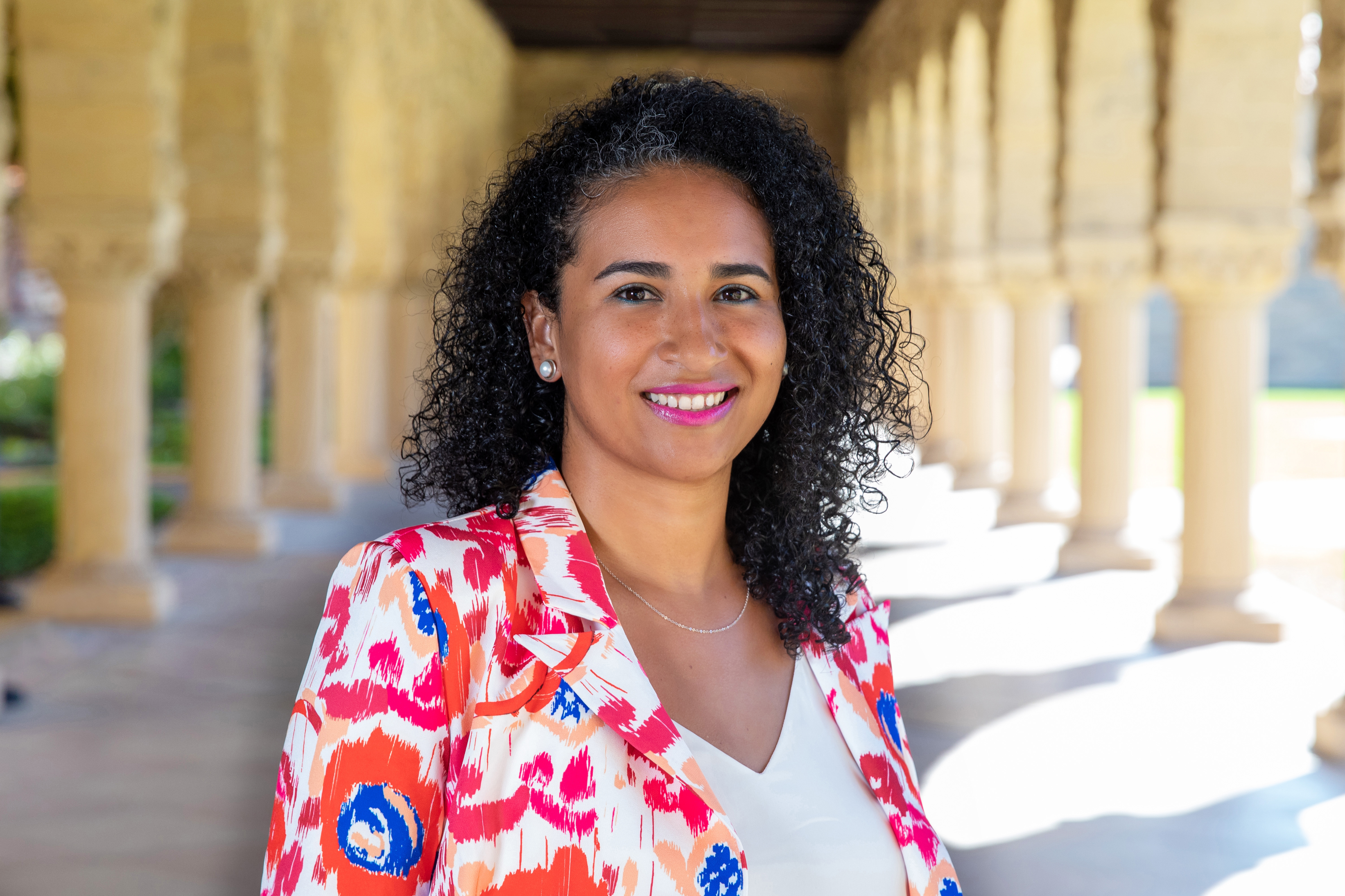 Headshot of a woman with dark curly hair wearing a printed pink and red blazer