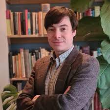 A man with short brown hair wearing a button down shirt and brown jacket stands against a bookshelf folding his arms
