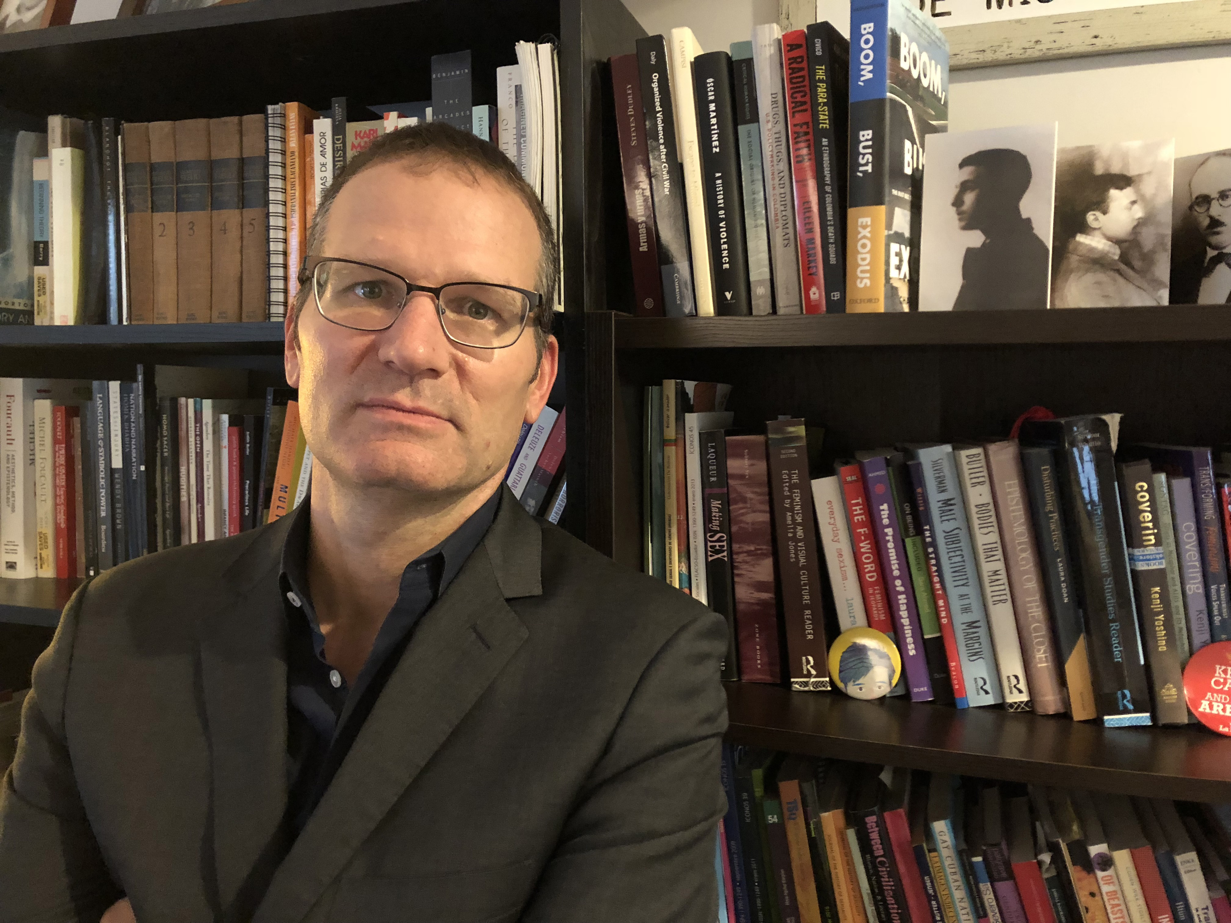 A man wearing glasses and a grey blazer against a bookshelf