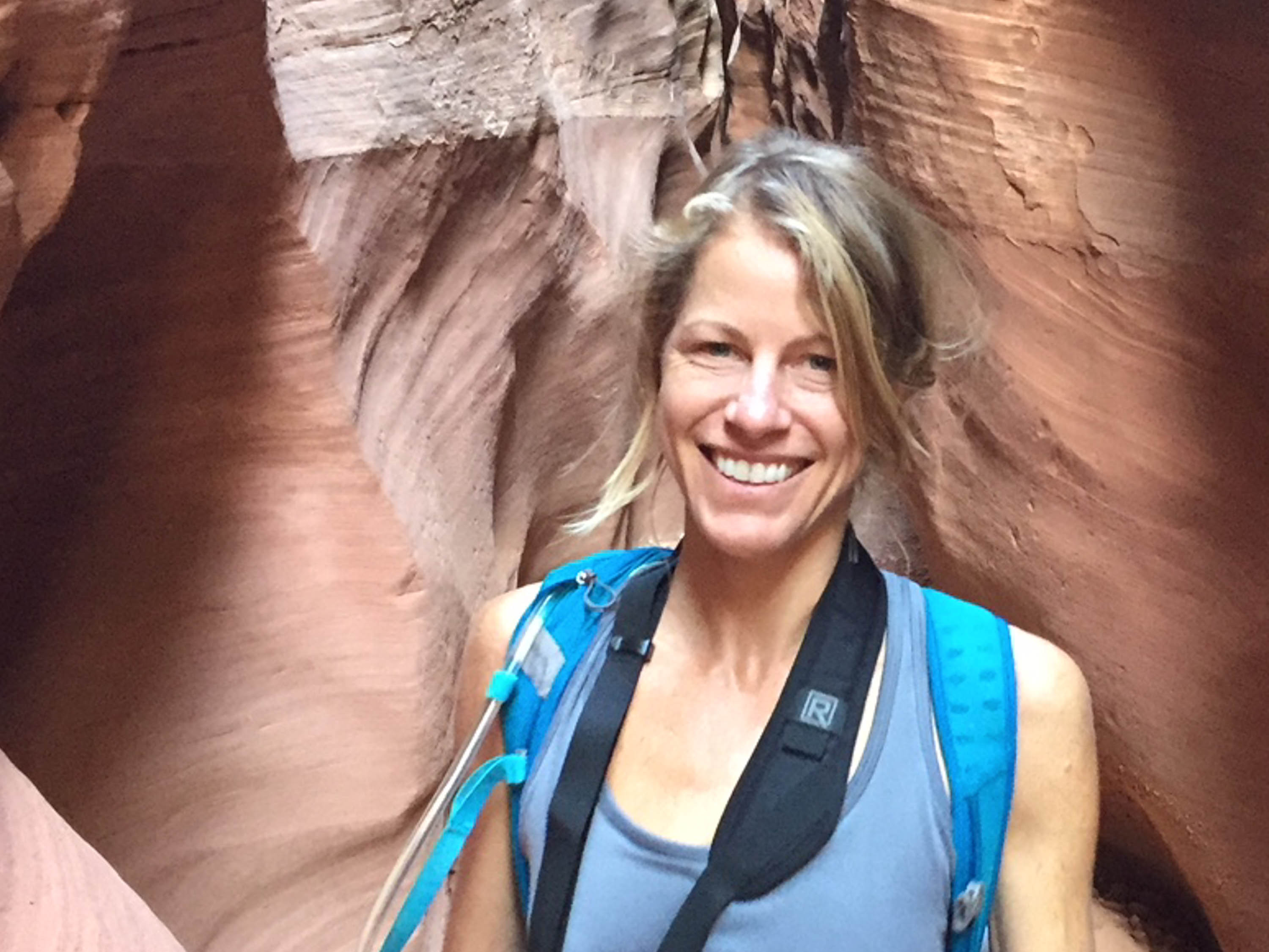 Carlyle wears a blue shirt and backpack while walking through a rocky formation