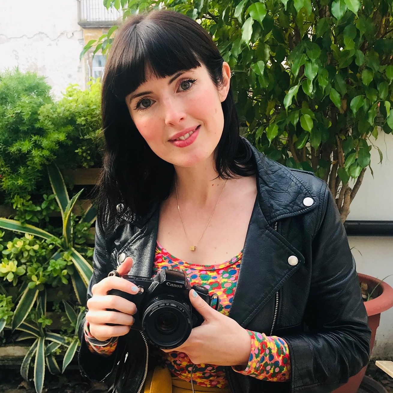 A woman with short dark hair and bangs wears a leather jacket and holds a camera