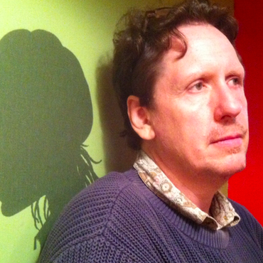 Trey wearing a blue sweater stands against a wall surrounded by green and red lighting