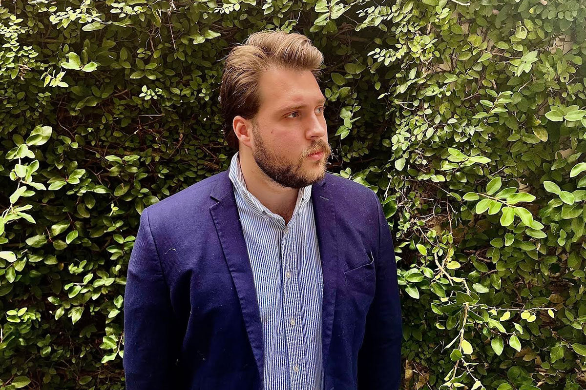 A man wearing a blue jacket and button down stands against a leafy background
