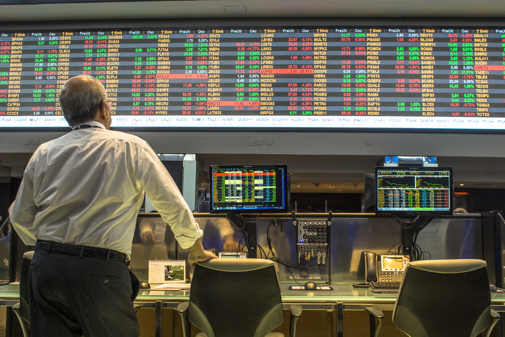 A man leans on his chair as he observes a large screen on the wall above with a string of red and green stock market values