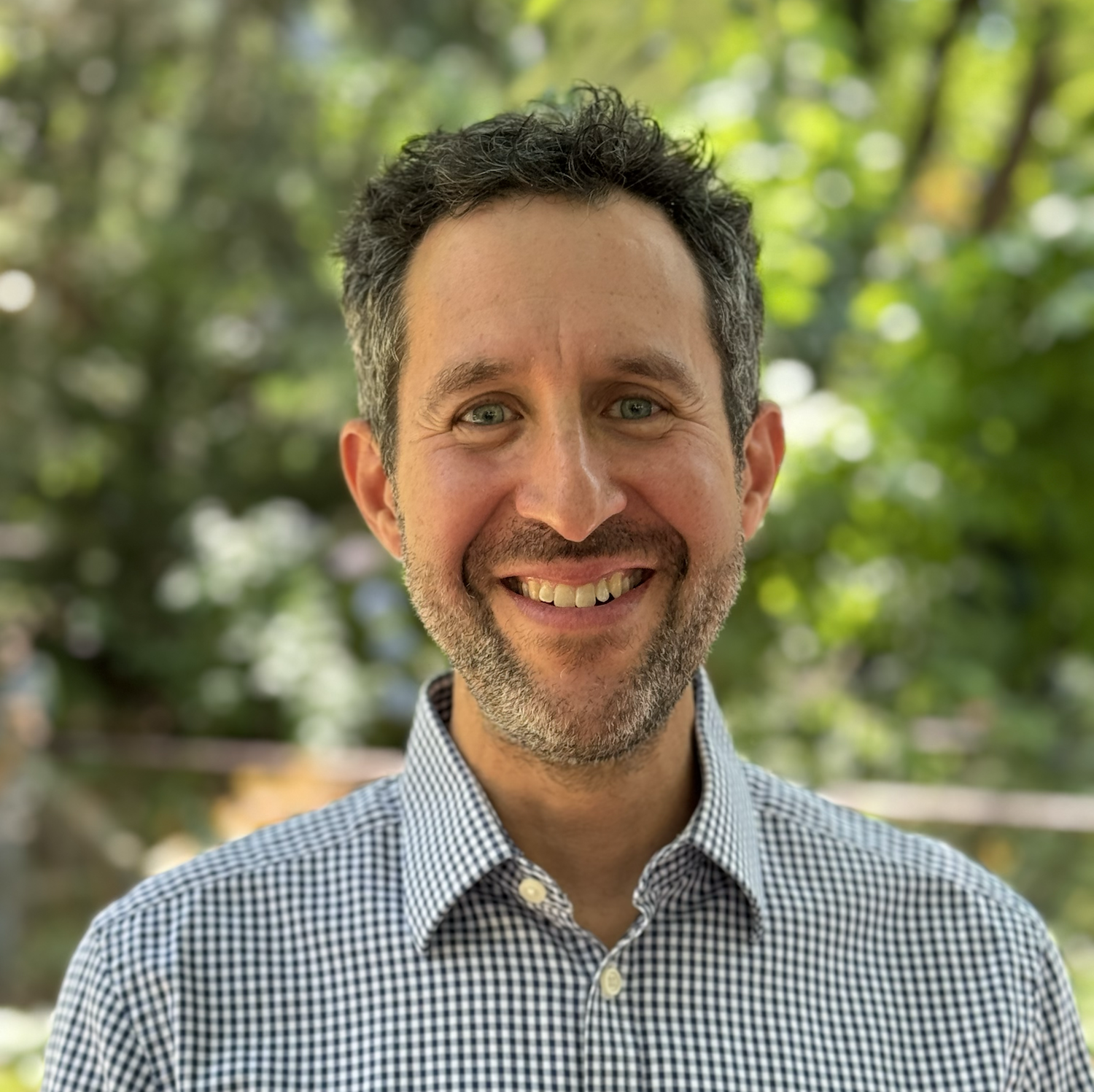 Headshot of man smiling in blue dress shirt with trees in background