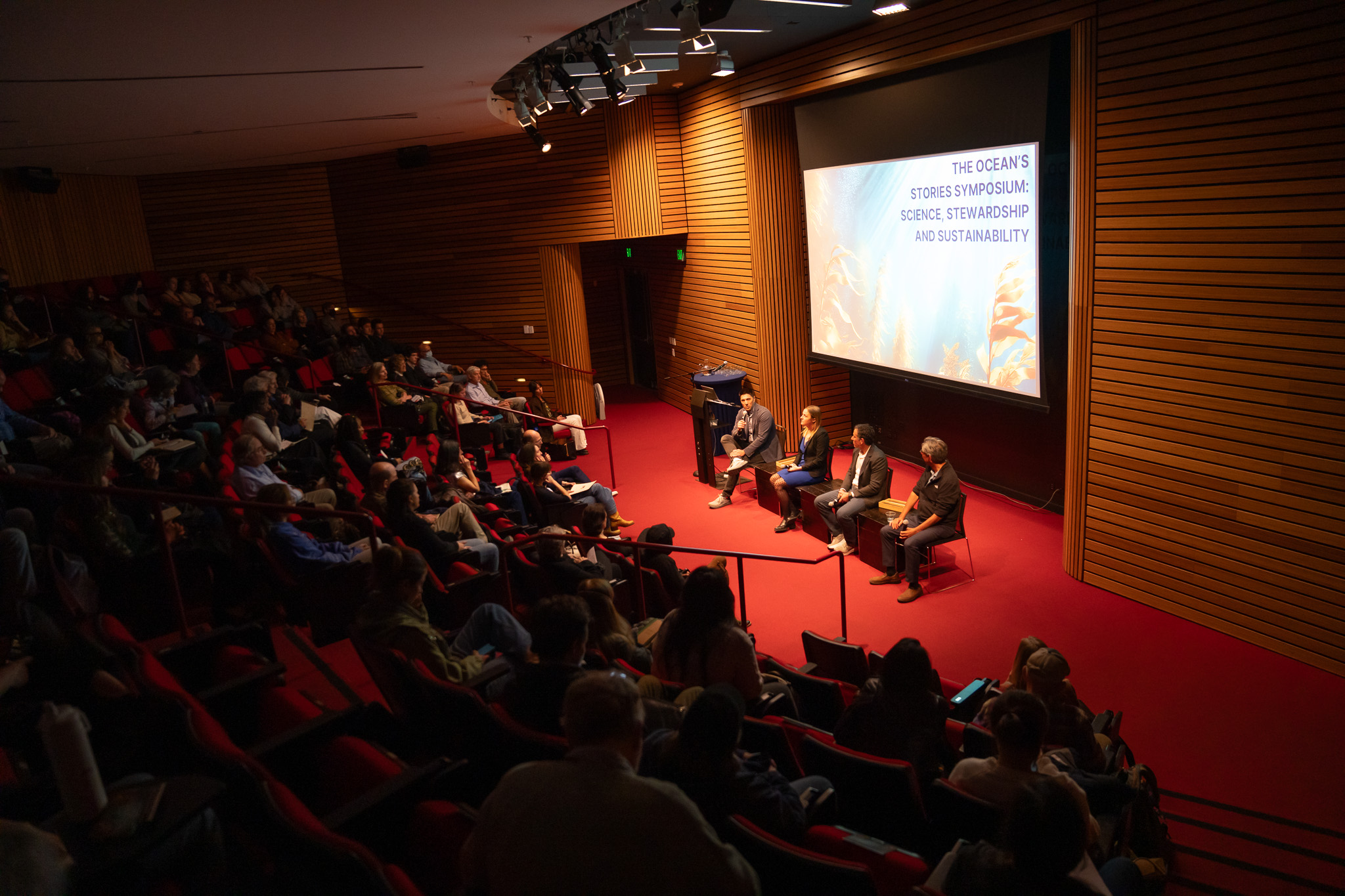 Four man sit for a panel in front of screen in auditorium