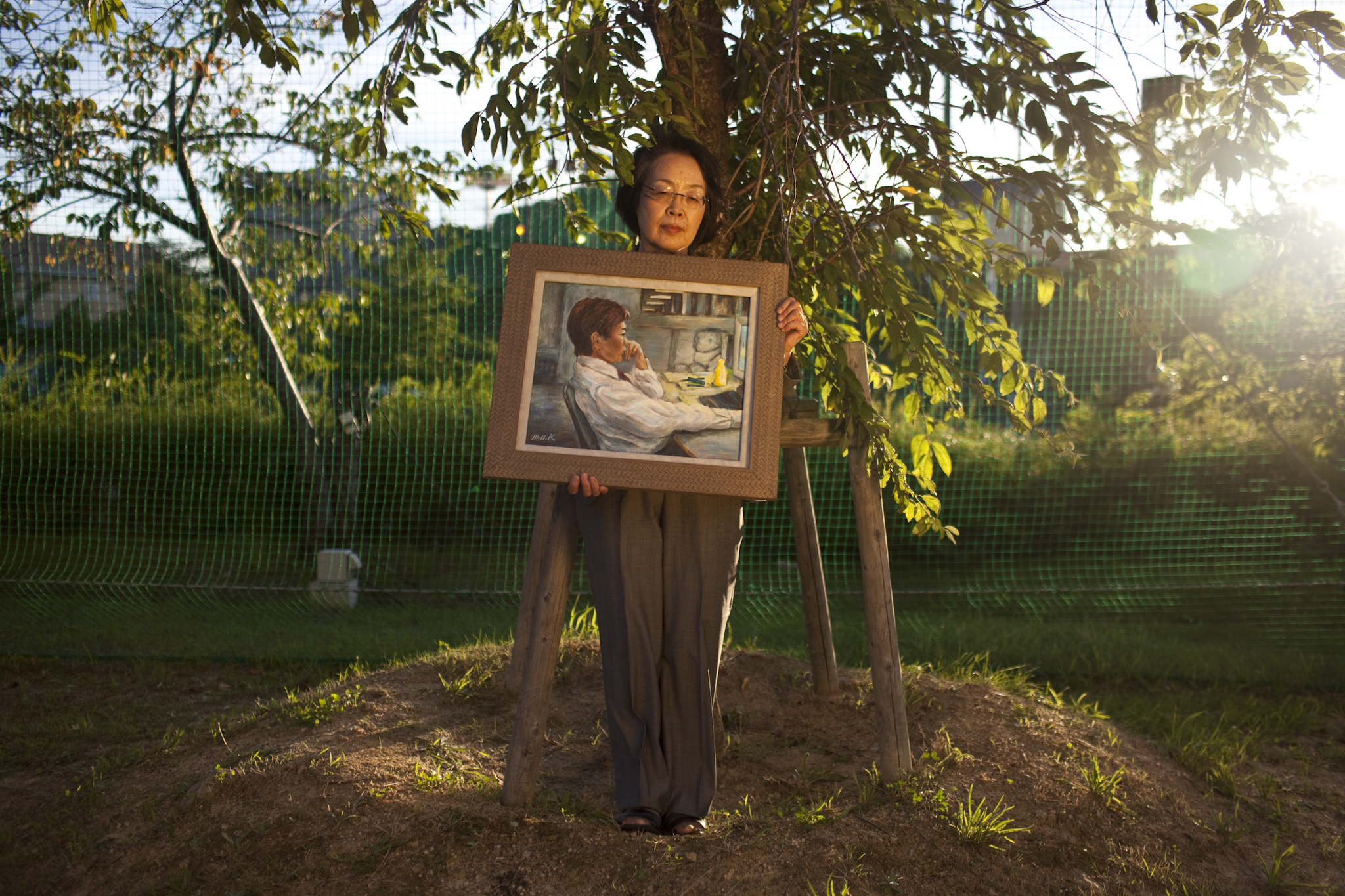 Michiyo Nishigaki, 67, with a painting of her son, poses under the cherry tree she planted after his death. Her son Naoya, a systems engineer who suffered from depression brought on by excessive overwork, killed himself in 2006. Image by Shiho Fukada. Japan, 2012. Media file: Nishigaki137.jpg