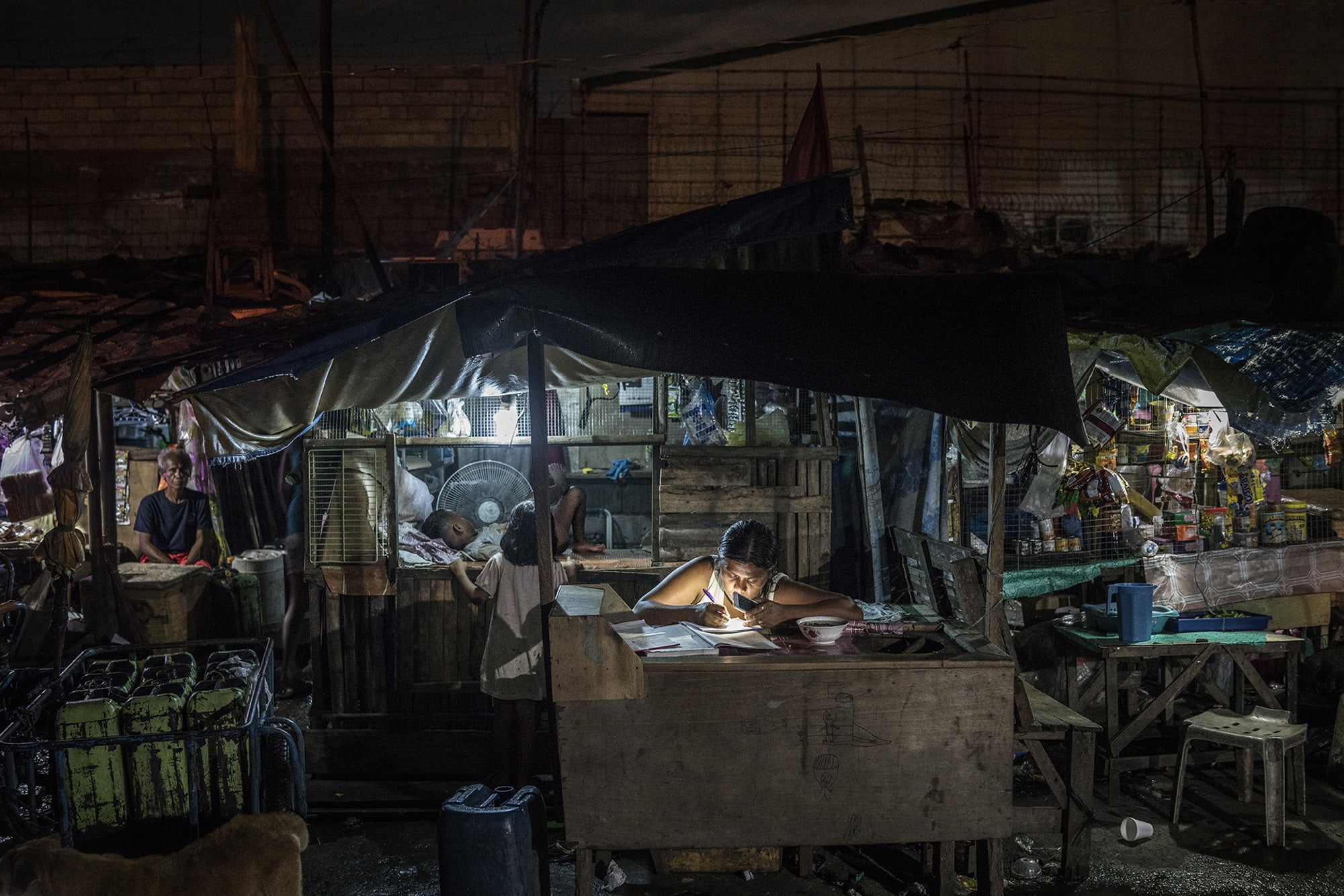 A woman works on bookkeeping using the light of her phone in the Market 3 slum in the Navotas Fish Port Complex. Market 3 has electricity but no plumbing. Residents often turn to drugs to alleviate hunger and stay awake for the long hours they work at low pay. Image by James Whitlow Delano. Philippines, 2017. A woman works on bookkeeping using the light of her phone in the Market 3 slum in the Navotas Fish Port Complex. Market 3 has electricity but no plumbing. Residents often turn to drugs to alleviate hunger and stay awake for the long hours they work at low pay. Image by James Whitlow Delano. Philippines, 2017.