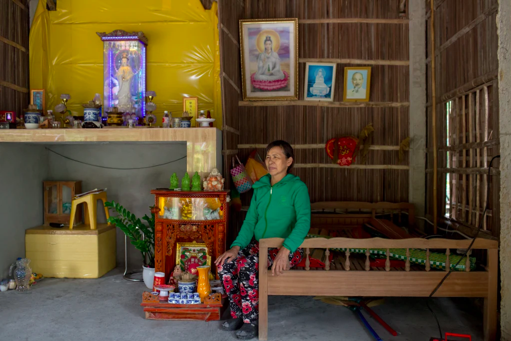 a woman wearing a green hoodie and floral patterned pants sits on a bed in her home made from palms