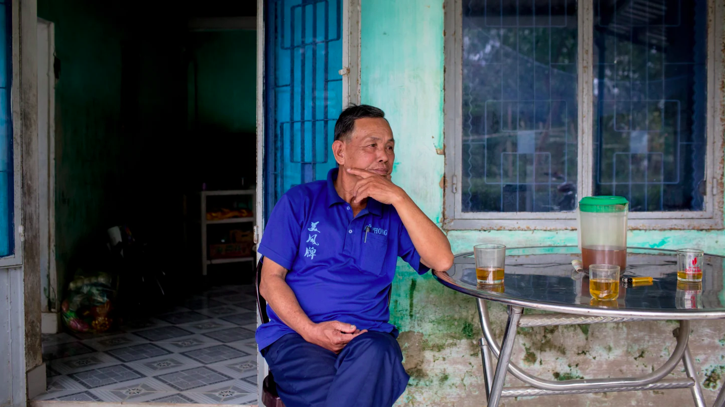 a man wearing a blue polo sits at a table outside with one hand in his lap and his elbow propped on the table with his hand on his chin