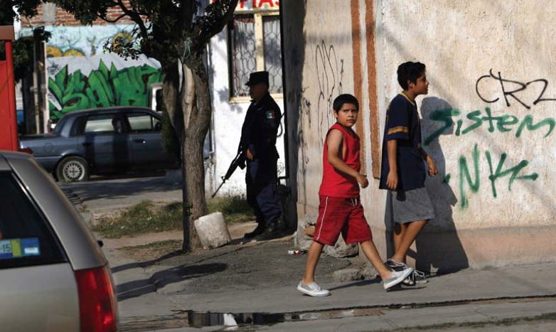 Boys walk by an armed official in Celaya in the Mexican state of Guanajuato, near the place where two grenades were hurled at a police outpost in November 2009. No one was hurt. Celaya is much more dangerous than Queretaro, which has a reputation for safety and is only 25 minutes away by car. Some say the difference is that drug cartels have agreed to keep Queretaro safe. Image by Alan Spearman, The Commercial Appeal.