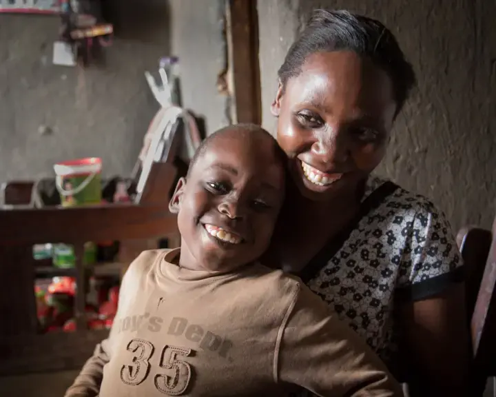 Prossy's son, Steven, was treated for infant hydrocephalus and spina bifida at the CURE Children's Hospital in Mbale, Uganda, several years ago. Steven can't walk and has outgrown his crutches, so his mother has to carry him. Prossy, a hairdresser, supports her three children alone since her husband abandoned the family. Image by Bridget Huber. Uganda, 2014. Media file: uganda_s-0143.jpg