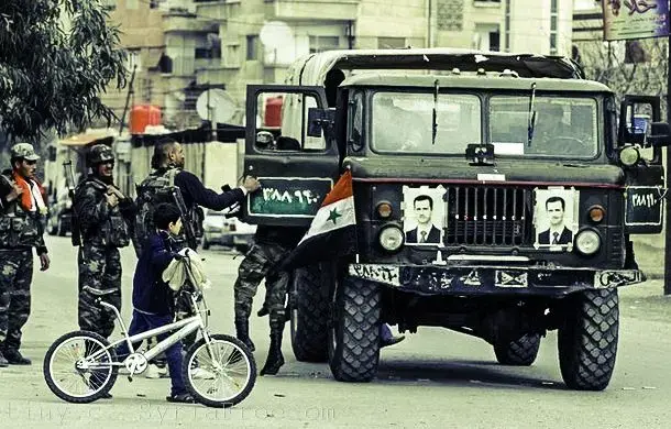 A young boy walks with a bicycle past Syrian army soldiers in the city of Harasta, 9 km north of Damascus. Photo courtesy of Freedom House flickr. Syria, 2012.
