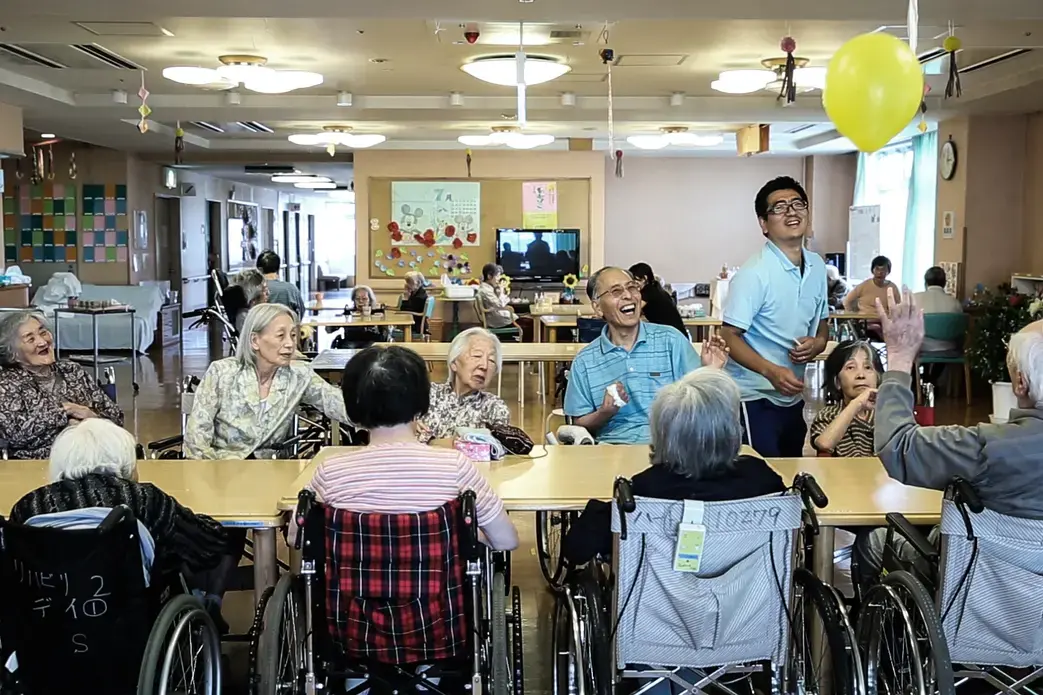 Seniors play with a baloon at the Flos Higashi-kojiya senior care facility in Tokyo. Image by Shiho Fukada. Japan, 2018. Media file: still001.jpg
