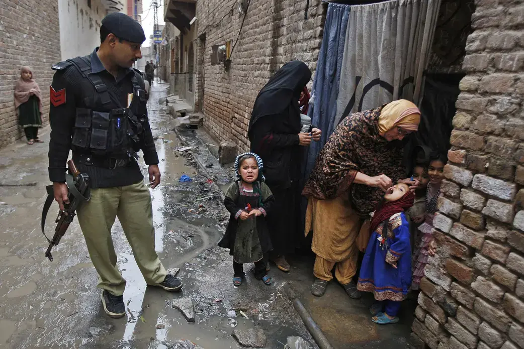 A Pakistani health worker, accompanied by an armed security guard, administers polio vaccine to a child in Peshawar in February. The day before, elsewhere in the same province, gunmen attacked a polio vaccination team, wounding a health worker and killing the driver. Image by Mohammad Sajjad, AP. Pakistan, 2014. Media file: polio3_natgeo-pakistan-06.adapt_.1190.2.jpg
