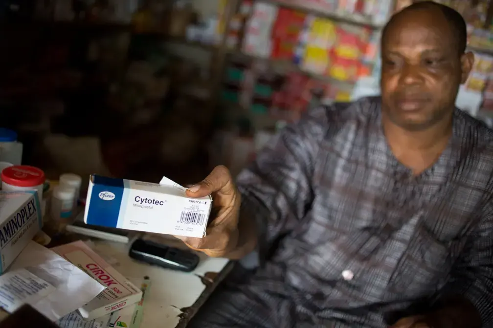 A vendor shows a customer the prescription drug Cytotec at a pharmacy in the Kirikiri neighborhood of Lagos. Cytotec, a trade name for the drug misoprostal, is used at lower doses to induce labor and slow post-partum bleeding, but has been widely used for self-induced abortions in Nigeria. The ten pills needed to induce abortion were sold to the customer — without a prescription — for 1,100 Naira, about $7. Image by Allison Shelley. Nigeria, 2013. Media file: nigeria_misoprostol_web002.jpg