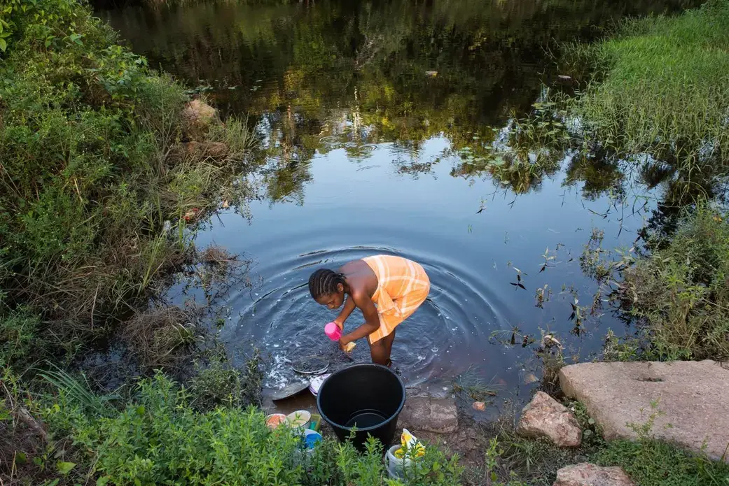 Osyanda Misidjan, 17, washes dishes on the banks of the Coermotibo River in her village of Adjuma Kondre, Suriname. Once surrounded by rainforest, the green palm trees and valley soccer field of the village are now surrounded by stony, bauxite-red plains left over from Alcoa's mining operations that ceased in 2015. Image by Stephanie Strasburg. Suriname, 2017.