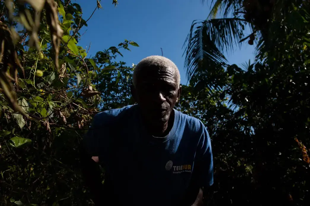 Nelson Adose Martines, 59, emerges from harvesting a cassava root by his family's shacks off of Suriname's Afobaka Road. Martines is part of the generations of Saamaka who still feel the effects of a 1964 government resettlement that moved thousands of residents from villages inundated by Alcoa's Brokopondo Reservoir. "We live off farming, farming is how we eat, how we live," said Martines. "But because a lot of villages relocated, we had to share these resources with more people." Image by Stephanie Strasburg. Suriname, 2017. Nelson Adose Martines, 59, emerges from harvesting a cassava root by his family's shacks off of Suriname's Afobaka Road. Martines is part of the generations of Saamaka who still feel the effects of a 1964 government resettlement that moved thousands of residents from villages inundated by Alcoa's Brokopondo Reservoir. "We live off farming, farming is how we eat, how we live," said Martines. "But because a lot of villages relocated, we had to share these resources with more people." Image by Stephanie…