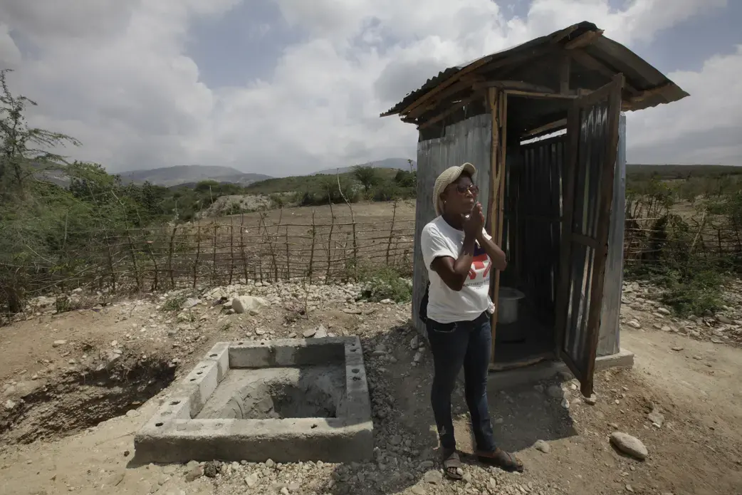 Public health worker Tamarra Joseph stands by a latrine that villagers built with help from the Community Health Initiative. When the latrine is full, the structure will be moved to the neighboring pit. The filled pit will be covered, and its contents will be composted for eventual use as fertilizer. Image by Mary Chind. Haiti, 2013. Media file: tamarra_joseph.jpg