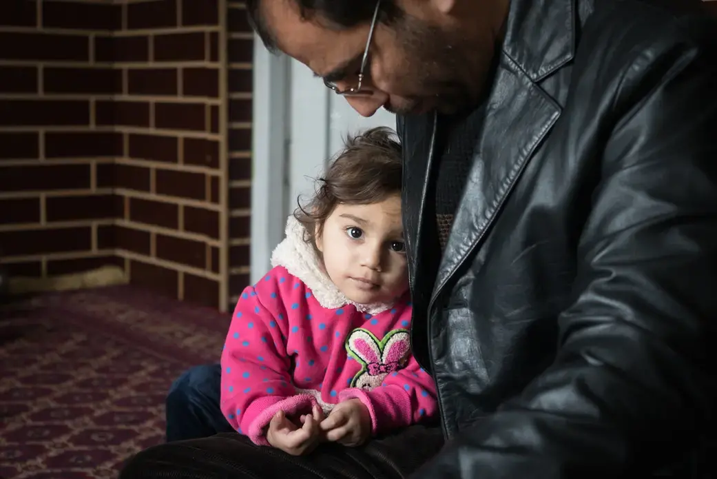 Dr. Wahid Qaderi holds his daughter in his home in Kabul, Afghanistan. His wife, mother, mother-in-law and five children recently crossed by boat from Turkey to Greece, while he stayed behind to try and sell their home. Image by Alexandria Bombach. Afghanistan, 2016. Media file: 1-howwechoose-alexandriabombach.jpg