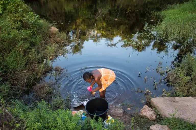 Osyanda Misidjan, 17, washes dishes on the banks of the Coermotibo River in her village of Adjuma Kondre, Suriname. Once surrounded by rainforest, the green palm trees and valley soccer field of the village are now surrounded by stony, bauxite-red plains left over from Alcoa's mining operations that ceased in 2015. Image by Stephanie Strasburg. Suriname, 2017.