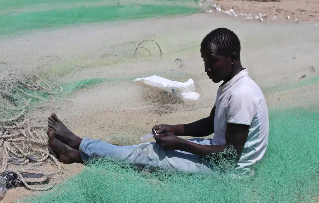 A fisherman mends his net on the beach at Gunjur fishing village. Image by Nosmot Gbadamosi. The Gambia, 2018. A fisherman mends his net on the beach at Gunjur fishing village. Image by Nosmot Gbadamosi. The Gambia, 2018.