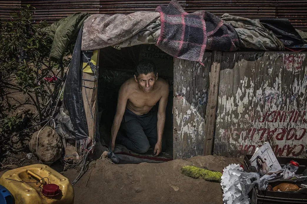 Luis, a deportee from the Mexican State of Querétaro, emerges from his makeshift shelter high atop the first desolate hilltop mesa east of the Pacific Ocean beside US/Mexico border wall. Image by James Whitlow Delano. Mexico, 2017. Luis—a deportee from the Mexican State of Querétaro, emerges from his makeshift shelter high atop the first desolate hilltop mesa east of the Pacific Ocean beside the U.S./Mexico border wall. Image by James Whitlow Delano. Mexico, 2017.