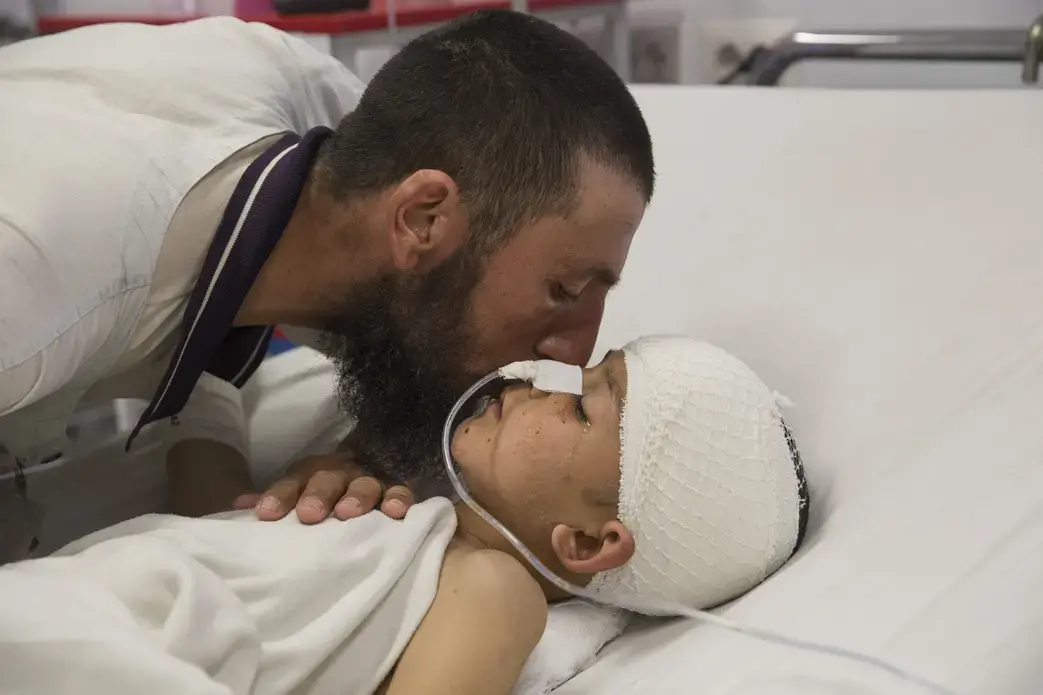 Abdul Kabir kisses his son, Noor Ahmad, 8, who is unconscious with a severe brain injury from an improvised explosive device (shell injury) from Mazar-E-Sharif Kabul on March 12, 2016. His father was also injured but recovered. Image by Paula Bronstein. Afghanistan, 2016. Abdul Kabir kisses his son, Noor Ahmad, 8, who is unconscious with a severe brain injury from an improvised explosive device (shell injury) from Mazar-E-Sharif Kabul on March 12, 2016. His father was also injured but recovered. Image by Paula Bronstein. Afghanistan, 2016.