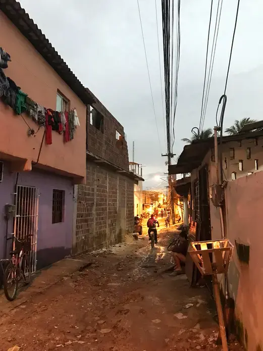Neighborhood on the outskirts of Recife, Pernambuco, where Mylene Helena dos Santos Ferreira, 23, lives with her partner and three sons. Her third child, Davi, was born in August 2015 with microcephaly and congenital Zika syndrome. Image by Poonam Daryani. Brazil, 2017. Image of a narrow street at dusk in a community on the outskirts of Recife, Pernambuco. There is an unpaved dirt road with small homes packed close together on either side. A teenager sits on a stoop in the shadows, while a young boy can be seen riding his bike down the street. Image by Poonam Daryani. Brazil, 2017.