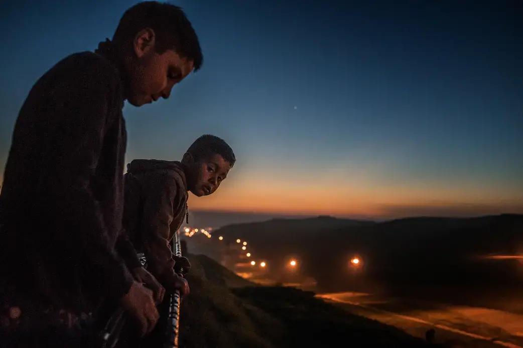 Young boys from the Arias family peer over the first, older US-built border wall at dusk into the flood-lit no-man's land created between the two walls/fences where US Border Patrol is present 24 hours a day, seven days a week.  Colonia Libertad, Tijuana, Mexico.  The Arias family is led by Estel, these boys’ grandmother and matriarch of the clan.  Their house actually rests up against the border wall and yet the family expressed no fear from human or drug traffickers.  Still their home, despite being…